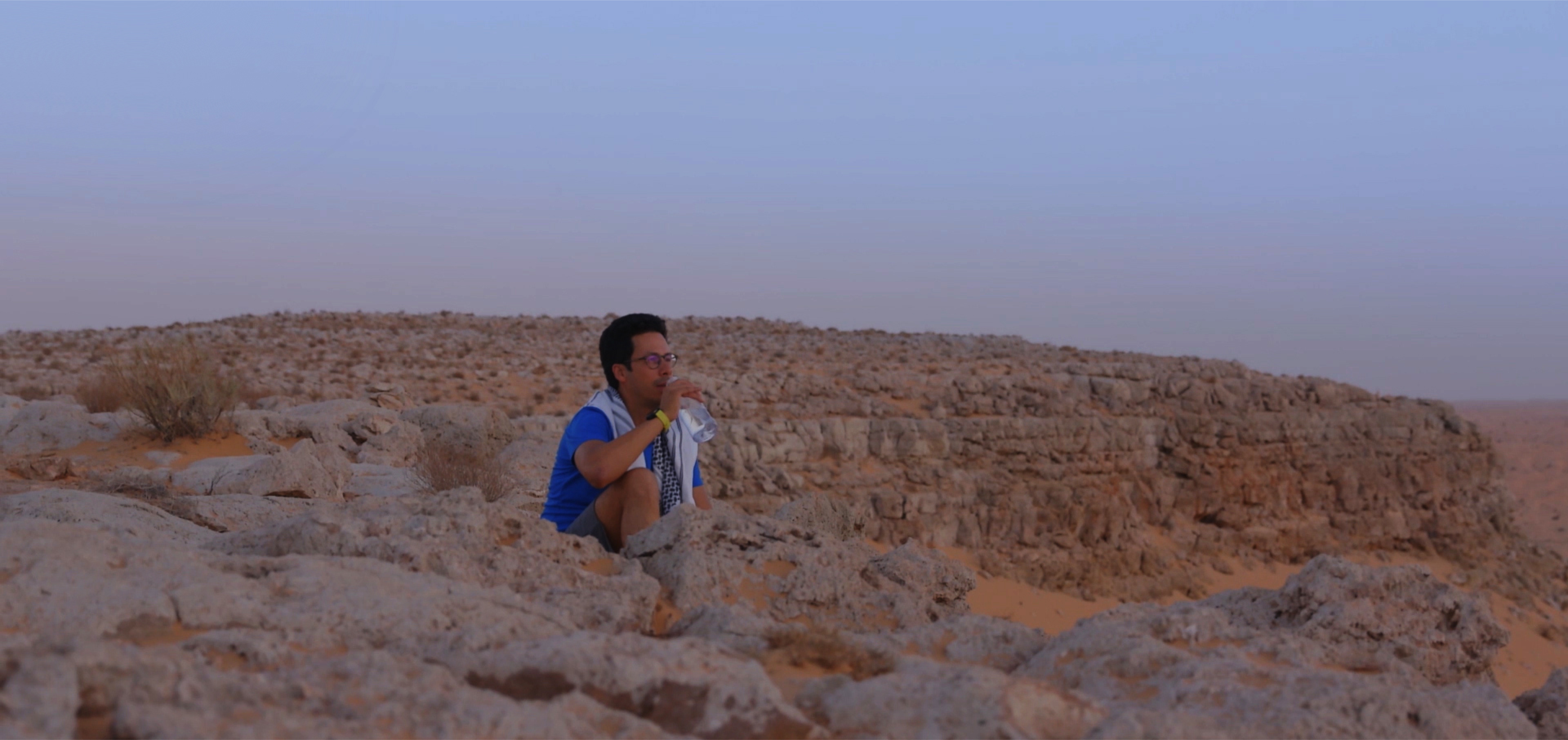Hedi sitting in a rocky part of the desert, drinking water looking atsunrise.